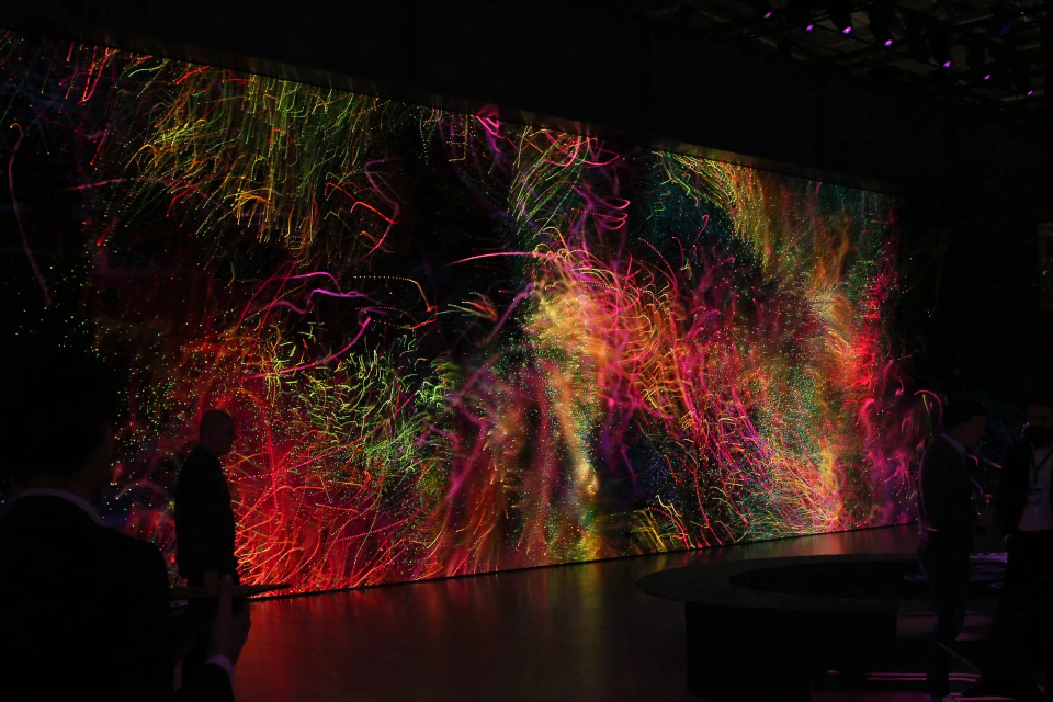 Multicolored generative particle trails spread across the darkened booth, visitors silhouetted in front of the LED wall