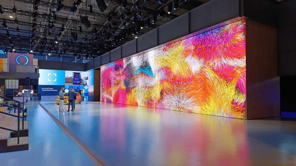The LED wall seen from across the exhibition hall, its warm generative composition visible from the neighboring booths